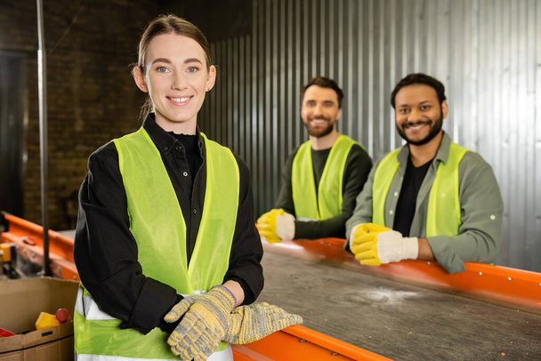 Smiling sorter in protective clothes and gloves looking at camera while standing near conveyor and blurred multiethnic colleagues in garbage sorting center, garbage sorting and recycling concept - Photo, Image