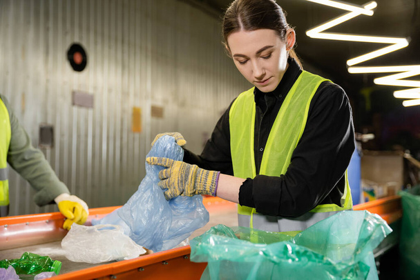 Joven trabajador en guantes y ropa protectora sosteniendo basura cerca de bolsa de plástico y transportador mientras trabaja en la estación de eliminación de residuos, clasificación de basura y el concepto de reciclaje - Foto, imagen