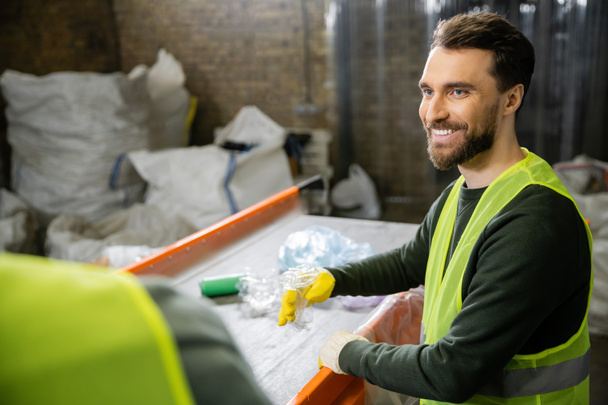 Cheerful male sorter in protective vest and gloves looking at blurred colleague while sorting garbage near conveyor in waste disposal station, garbage sorting and recycling concept - Photo, Image