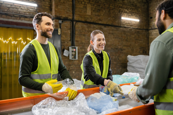 Cheerful sorters in vests and protective gloves sorting garbage and looking at blurred indian colleague near conveyor in waste disposal station, garbage sorting and recycling concept - Photo, Image