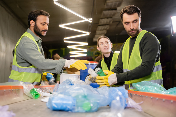 Wieloetniczne sortowniki w ubraniach ochronnych i rękawiczkach rozmawiające z kolegą podczas pracy z plastikowymi odpadami na przenośniku w stacji utylizacji odpadów, koncepcja sortowania i recyklingu odpadów - Zdjęcie, obraz