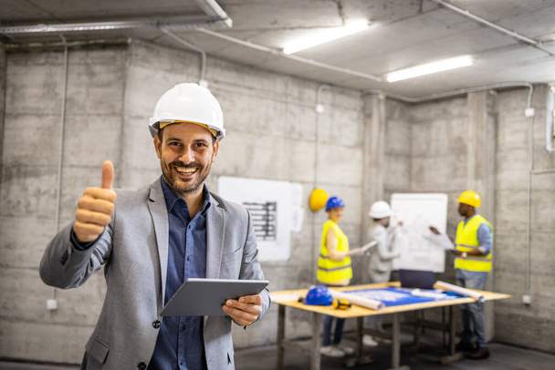 Portrait of structural engineer in business suit and hardhat holding thumb up at construction site. - Photo, Image