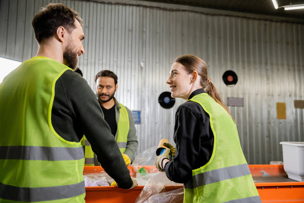 Smiling woman in protective uniform and gloves holding garbage and talking to interracial colleagues while working together near conveyor in waste disposal station, garbage sorting concept - Photo, Image