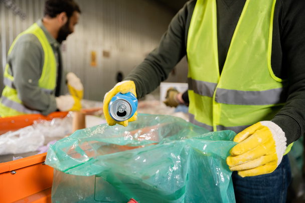 Cropped view of man in protective vest and gloves putting tin can in plastic bag while working near blurred multiethnic colleagues and conveyor in waste disposal station, recycling concept - Photo, Image