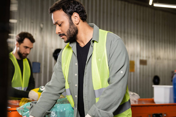 Bearded indian worker in high visibility vest separating trash while working near blurred conveyor and colleague in waste disposal station at background, recycling concept - Photo, Image