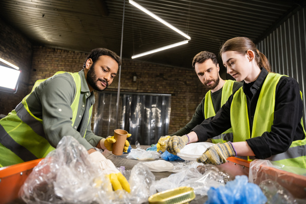 Multiethnic workers in protective gloves and vests separating and taking trash from conveyor while working together in blurred waste disposal station, recycling concept - Photo, Image