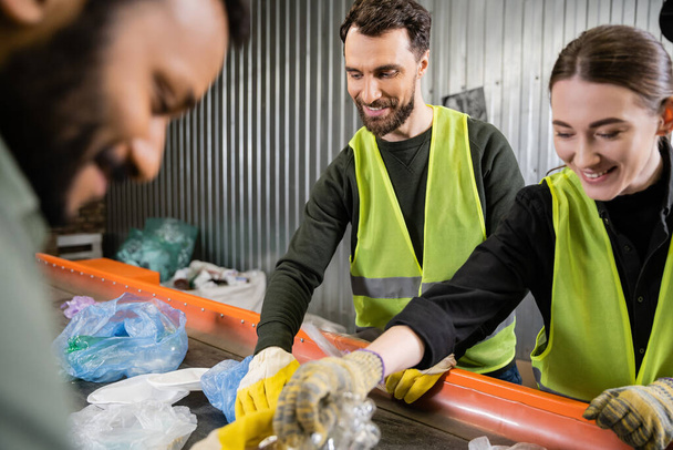Uśmiechnięty i brodaty pracownik w kamizelce ochronnej i rękawiczkach zabierający i oddzielający śmieci na przenośniku w pobliżu wesołych międzyrasowych kolegów pracujących w sortowni śmieci, koncepcja recyklingu - Zdjęcie, obraz