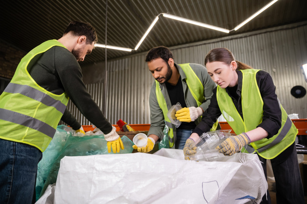 Young worker in reflective vest and gloves holding plastic containers near sack while sorting trash with multiethnic colleagues in waste disposal station, garbage sorting and recycling concept - Photo, Image