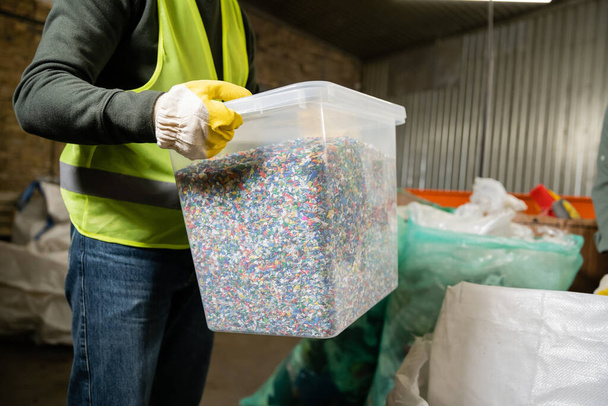 Обрезанный вид работника в жилете и контейнере для хранения перчаток с пластиком для переработки во время работы в центре сортировки мусора, концепции сортировки и переработки мусора - Фото, изображение