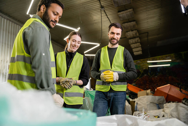 Trabajadores multiétnicos sonrientes con chalecos y guantes protectores que miran sacos borrosos con basura para reciclar mientras trabajan juntos en la estación de eliminación de residuos, concepto de reciclaje - Foto, Imagen