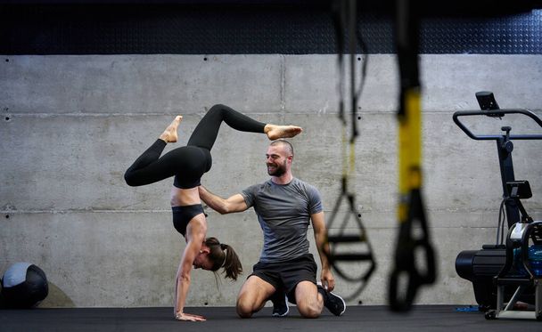 A muscular man assisting a fit woman in a modern gym as they engage in various body exercises and muscle stretches, showcasing their dedication to fitness and benefiting from teamwork and support. - 写真・画像