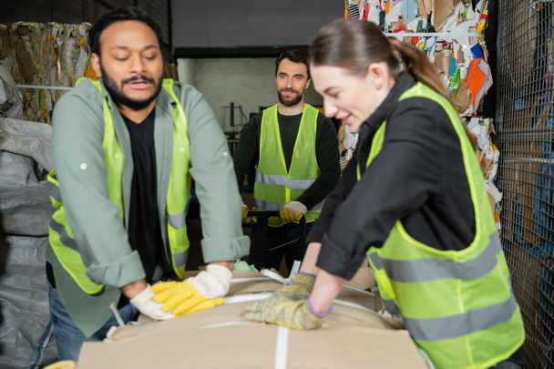 Smiling worker in reflective vest and gloves standing near hand pallet truck while blurred interracial colleagues pressing waste paper in garbage sorting center, waste sorting and recycling concept - Photo, Image