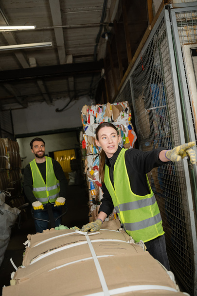 worker in protective vest and gloves pointing with finger away while standing near waste paper and colleague with hand palette truck in garbage sorting center, waste sorting and recycling concept - Photo, Image