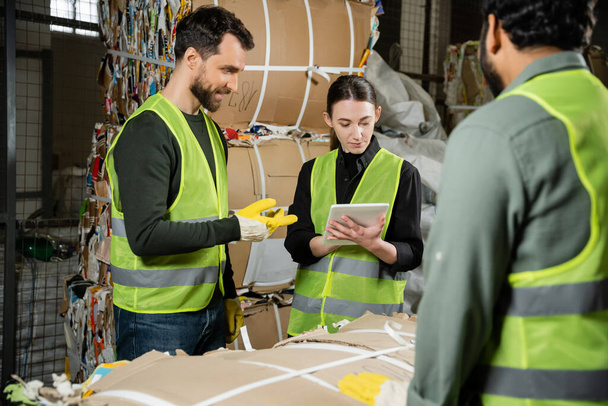 Young worker in protective vest and gloves using digital tablet near smiling multiethnic colleagues and waste paper in garbage sorting center, waste sorting and recycling concept - Photo, Image