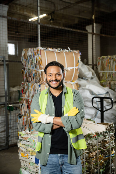 Smiling and bearded worker in protective gloves and vest crossing arms and looking at camera while standing near blurred waste paper in waste disposal station, garbage sorting and recycling concept - Photo, Image