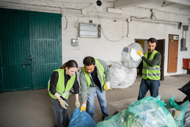 Trabajadores multiétnicos en chalecos y guantes de protección que trabajan con bolsas de plástico cerca de su colega en la estación de eliminación de residuos, clasificación de basura y el concepto de reciclaje - Foto, Imagen