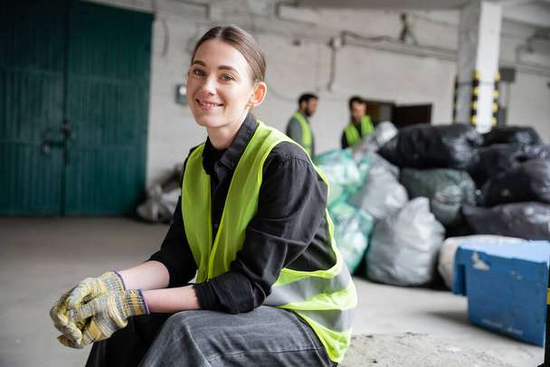 Cheerful young worker in safety vest and gloves looking at camera while resting and sitting near blurred plastic bags in garbage sorting center, recycling concept - Photo, Image