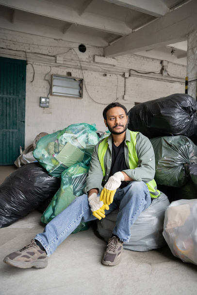 Trabajador indio masculino en chaleco de alta visibilidad y guantes protectores sentado en bolsas de plástico con basura mientras trabaja en el centro de clasificación de basura, concepto de reciclaje - Foto, Imagen