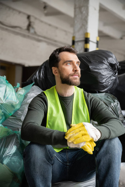 Trabajador barbudo en chaleco de alta visibilidad y guantes mirando hacia otro lado mientras está sentado cerca de bolsas de plástico con basura en el centro de clasificación de basura borrosa, concepto de reciclaje - Foto, Imagen