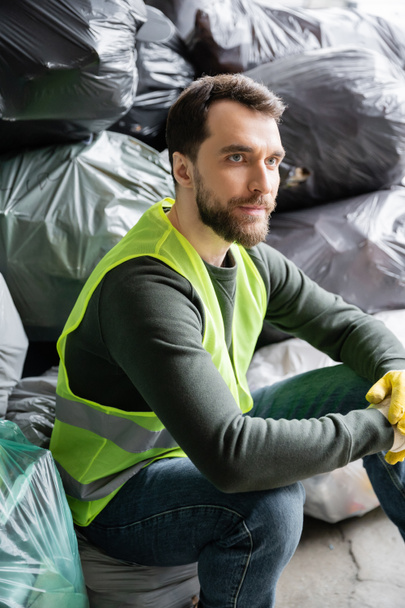 Trabajador barbudo con chaleco fluorescente y guantes mirando hacia otro lado mientras está sentado cerca de bolsas de plástico borrosas con basura en el centro de clasificación de basura, concepto de reciclaje - Foto, imagen