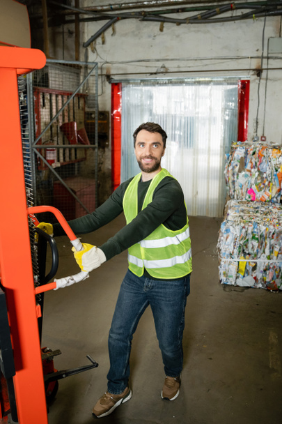 Trabajador sonriente en chaleco de seguridad y guantes tirando de la transpaleta de mano mientras trabaja cerca de papel de desecho borroso en el centro de clasificación de basura en segundo plano, concepto de reciclaje - Foto, Imagen