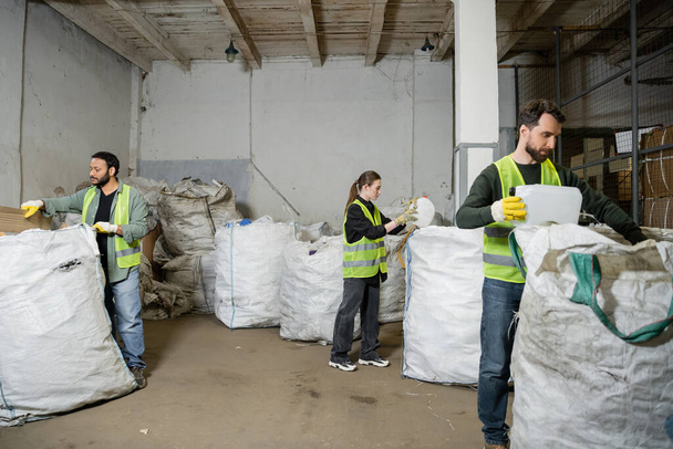 Lavoratori multietnici in giubbotti e guanti ad alta visibilità che separano i rifiuti nei sacchi mentre lavorano nella stazione di smaltimento dei rifiuti, nella raccolta differenziata dei rifiuti e nel concetto di riciclaggio - Foto, immagini