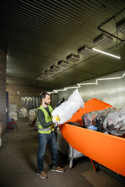 Πλευρική άποψη γενειοφόρου αρσενικού εργαζομένου σε γάντια και γιλέκο φθορισμού βάζοντας σάκο με σκουπίδια σε δοχείο σε θολή σταθμό διάθεσης αποβλήτων, διαλογή απορριμμάτων και έννοια ανακύκλωσης - Φωτογραφία, εικόνα