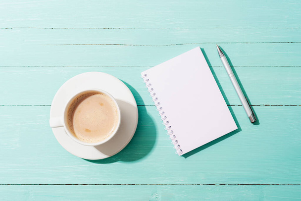 Una taza de café, bloc de notas y bolígrafo en la mesa azul turquesa a la luz del sol. Vista superior, plano, maqueta. - Foto, Imagen