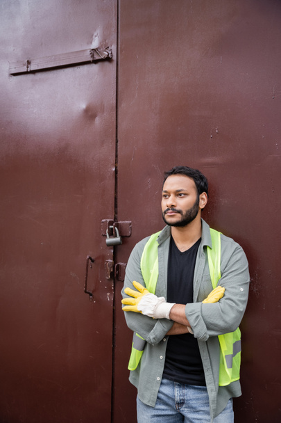 Der bärtige indische Arbeiter in Warnweste und Handschuhen kreuzt die Arme, während er in der Nähe der Tür der Mülldeponie steht, Müllsortier- und Recyclingkonzept - Foto, Bild