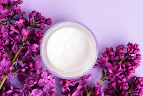 Cream jar and lilac flowers on light purple background. Natural cosmetics, skin care concept. Top view, flat lay. - Foto, Imagem