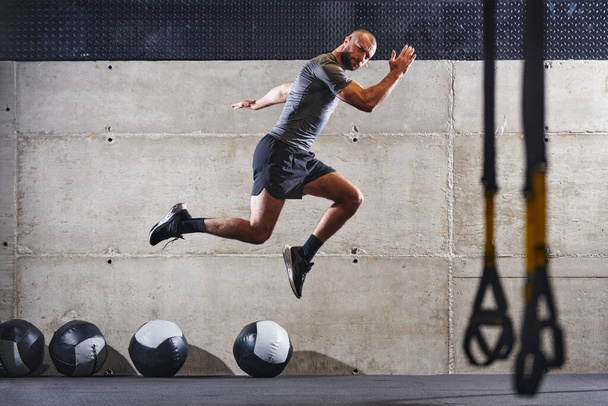 A muscular man captured in air as he jumps in a modern gym, showcasing his athleticism, power, and determination through a highintensity fitness routine. - Photo, image