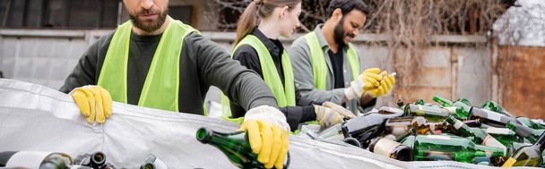 Bearded sorter in protective gloves and vest putting glass bottle in sack near blurred multiethnic colleagues in outdoor waste disposal station, garbage sorting and recycling concept, banner  - Photo, Image