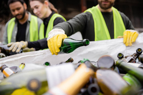 Blurred worker in protective vest and gloves putting glass bottle in sack while working with multiethnic colleagues outdoors in waste disposal station, garbage sorting and recycling concept - Photo, Image