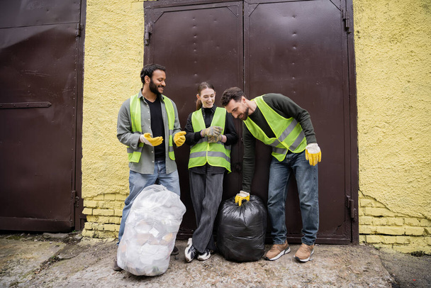 Travailleur joyeux en gilet et gants haute visibilité mettant sac poubelle près des collègues internationaux et porte de la station d'élimination des déchets à l'extérieur, concept de tri et de recyclage des déchets - Photo, image