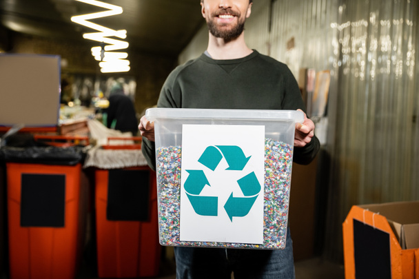 Vista recortada del voluntario sonriente y barbudo sosteniendo la papelera con el cartel de reciclaje en la estación de eliminación de residuos borrosa en el fondo, clasificación de la basura y el concepto de reciclaje - Foto, Imagen