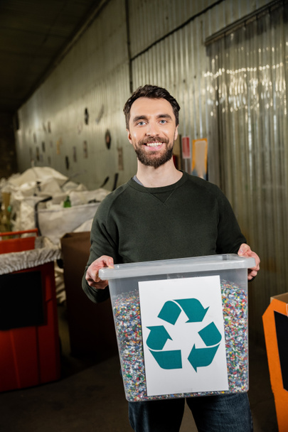 Χαρούμενος και γενειοφόρος εθελοντής που κοιτάζει την κάμερα ενώ κρατάει τον κάδο απορριμμάτων με την ένδειξη ανακύκλωσης στο σταθμό διάθεσης αποβλήτων στο παρασκήνιο, τη διαλογή και την έννοια της ανακύκλωσης - Φωτογραφία, εικόνα