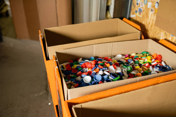 Scatole di cartone con tappi di plastica colorati per il riciclo in una stazione di smaltimento dei rifiuti sfocata sullo sfondo, la raccolta differenziata e il concetto di riciclaggio - Foto, immagini