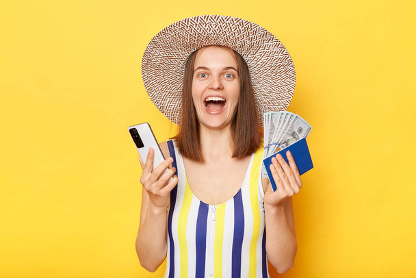 Overjoyed woman tourist holding cellphone tickets passport have excellent journey wearing striped swimming suit posing isolated over yellow background. - Фото, зображення