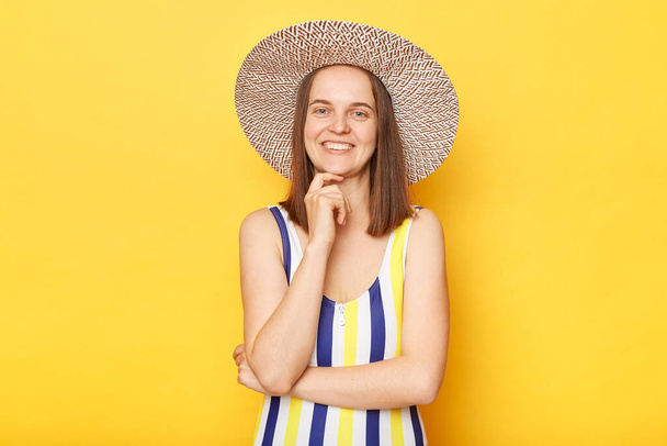 Pretty smiling happy woman wearing striped swimming suit and hat isolated on yellow background keeps hands under chin looking at camera with happy expression. - Foto, Imagem