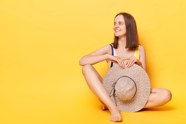 Confident attractive woman wearing striped swimming suit and hat isolated on yellow background sitting looking away at advertisement area, copy space for promotion. - Fotografie, Obrázek