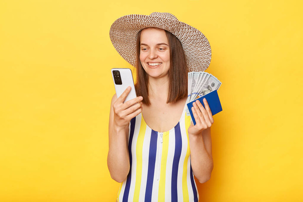 Optimistic smiling woman tourist holding cellphone passport booking tickets planning traveling wearing striped swimming suit posing isolated over yellow background - Φωτογραφία, εικόνα