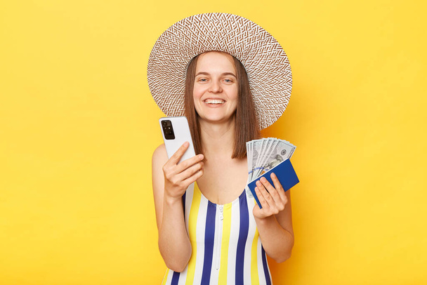 Smiling attractive woman tourist using mobile phone standing with tickets passport in hands traveling abroad wearing striped swimming suit posing isolated over yellow background - Photo, image