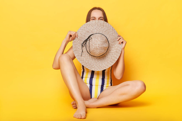 Shy smiling woman wearing striped swimming suit isolated on yellow background hiding her face with her hat summertime tropical vacation. - Фото, зображення