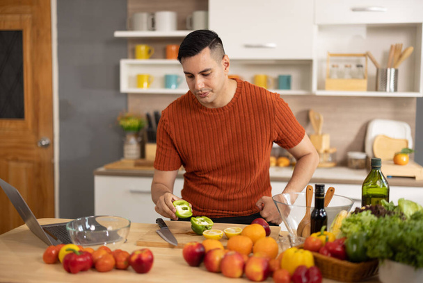 A homely and healthy male prepares nutritious diet, including variety of fruits. Following cooking show on laptop Mastery of peeling, chopping, and slicing fruits to prepare them in artistic style. - Photo, image