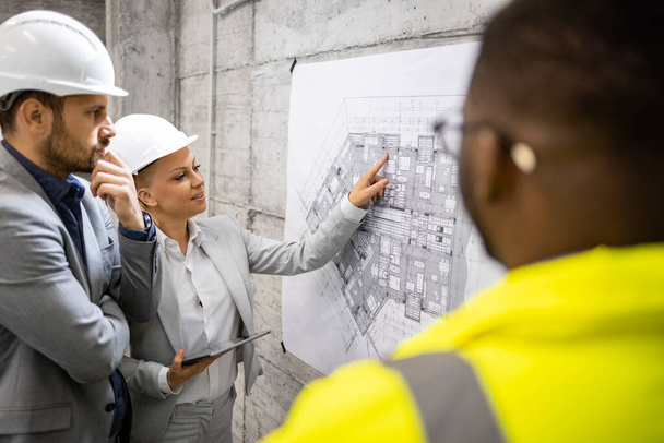 Rakennustyöntekijä ja arkkitehdit tapaavat rakennustyömaalla analysoimassa projektisuunnitelmaa ja rakennuksen seuraavaa vaihetta. - Valokuva, kuva
