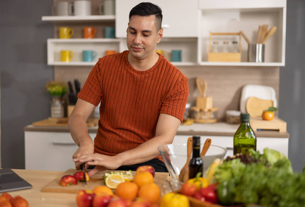 A homely and healthy male prepares nutritious diet, including variety of fruits. Following cooking show on laptop Mastery of peeling, chopping, and slicing fruits to prepare them in artistic style. - Photo, image