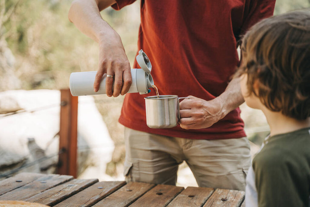 Father dad pours hot coffee tea from thermos into the mug on a family picnic in the mountains. Child school boy kid is watching his dad filling the cup with hot beverage. - 写真・画像