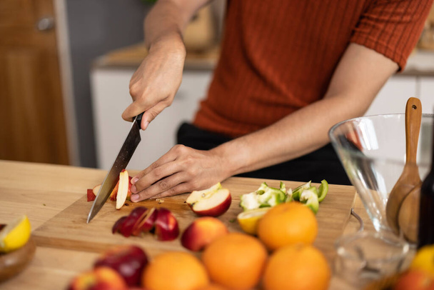 A homely and healthy male prepares nutritious diet, including variety of fruits. Following cooking show on laptop Mastery of peeling, chopping, and slicing fruits to prepare them in artistic style. - Foto, imagen