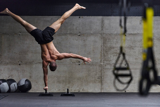 A muscular man in a handstand position, showcasing his exceptional balance and body control while performing a variety of exercises to enhance his overall body stability and strength in a modern gym. - Foto, Imagem