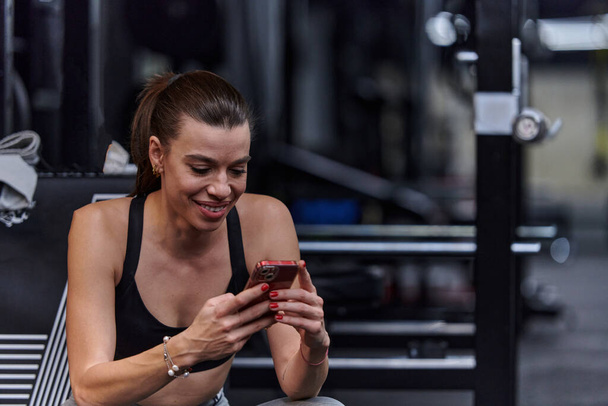 A fit woman in the gym taking a break from her training and uses her smartphone, embracing the convenience of technology to stay connected. - Photo, Image